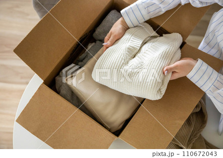 Woman hands putting warm neutral sweater into cardboard box seasonal storage method and donation 110672043