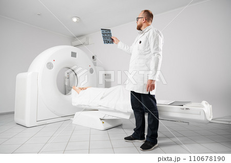 Medical computed tomography or MRI scanner. Doctor holding and examining results of MRI. Woman patient lying on CT scan bed. Concept of medicine, healthcare and modern diagnostics. Medical computed tomography or MRI scanner. Doctor holding and examining results of MRI. Woman patient lying on CT scan bed. Concept of medicine, healthcare and modern diagnostics. 110678190