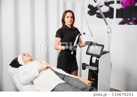 Female cosmetologist holding radiofrequency device and looking at camera while standing by client in cosmetology clinic. Woman receiving rejuvenation skincare treatment in beauty salon. Female cosmetologist holding radiofrequency device and looking at camera while standing by client in cosmetology clinic. Woman receiving rejuvenation skincare treatment in beauty salon. 110678201