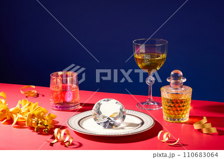 A glass, a cocktail glass and a glass bottle containing a clear yellow liquid, a diamond on a ceramic plate, surrounded by flowers and ribbons. Minimalist background 110683064