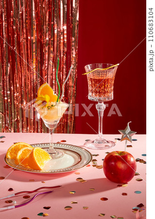 A cocktail and fresh orange slices on a ceramic plate, next to a glass of wine. Confetti, candles and red apples on a pink background. Sparkling strings hang on a dark red background. 110683104