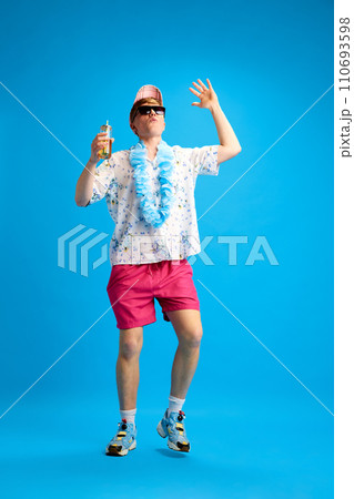 Young guy in casual stylish clothes, shrift and shorts, in sunglasses, drinking lemonade and dancing against blue studio background 110693598