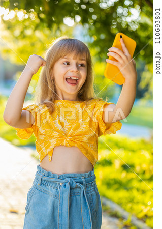 Preteen child girl use smartphone play online game celebrating win good message news in city park Preteen child girl use smartphone play online game celebrating win good message news in city park 110693801