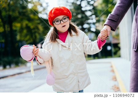Cute girl in eyeglasses wearing coat red beret with special needs holding mother's hand on a walk outdoor. Cute girl in eyeglasses wearing coat red beret with special needs holding mother's hand on a walk outdoor. 110694072