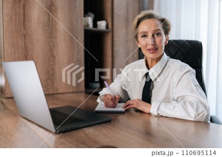 Successful and happy middle-aged business woman looking at camera sitting inside office working with documents. Portrait of satisfied woman writing in notebook and working using laptop at work. 110695114