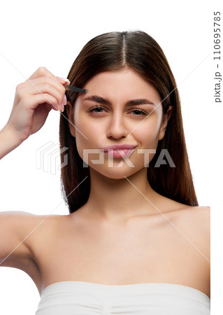 Young emotional girl with tweezing her eyebrows and looking at camera giving attention to beauty and facial grooming against white studio background. Young emotional girl with tweezing her eyebrows and looking at camera giving attention to beauty and facial grooming against white studio background. 110695785