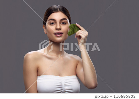 Portrait of young woman holding green gua sha stone near her face, portraying clean and natural beauty regimen. Portrait of young woman holding green gua sha stone near her face, portraying clean and natural beauty regimen. 110695805