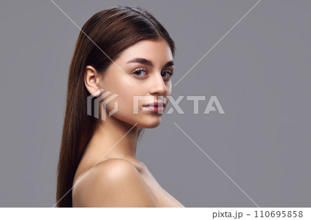 Portrait of young woman with clear skin, natural makeup, and neat hair looking over shoulder at camera against gray background. 110695858