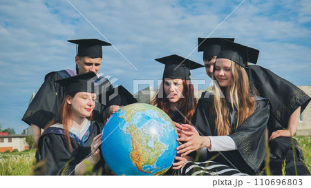 Graduates in black robes examine a geographical globe sitting on the grass. Graduates in black robes examine a geographical globe sitting on the grass. 110696803