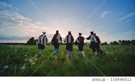 Six graduates in robes walk against the backdrop of the sunset. Six graduates in robes walk against the backdrop of the sunset. 110696812