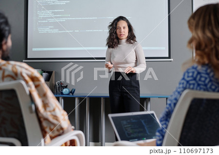 Serious female businesswoman talking to colleagues about code projected on digital board behind her 110697173