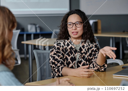 Young woman talking to her unrecognizable female colleague while sitting at desk in office 110697252