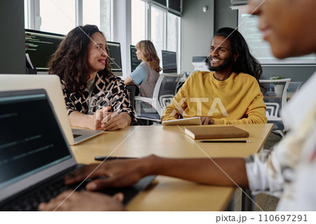 Biracial colleagues smiling at each other while having conversation at desk during workday in office Biracial colleagues smiling at each other while having conversation at desk during workday in office 110697291