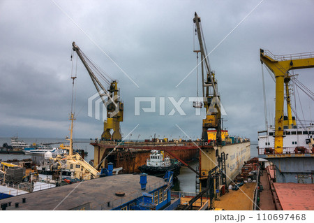 Industrial ship repair yard with floating dock, cranes hoist vessels for maintenance. Maritime engineering, dockyard activity under cloudy sky. Shipping industry, ship refitting, heavy machinery work. Industrial ship repair yard with floating dock, cranes hoist vessels for maintenance. Maritime engineering, dockyard activity under cloudy sky. Shipping industry, ship refitting, heavy machinery work. 110697468