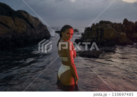 Attractive female model enjoys nighttime dip in sea illuminated by crimson lights, her style radiant in designer swimwear. The tranquil waters reflect her poised elegance amidst rocky seaside. Attractive female model enjoys nighttime dip in sea illuminated by crimson lights, her style radiant in designer swimwear. The tranquil waters reflect her poised elegance amidst rocky seaside. 110697485