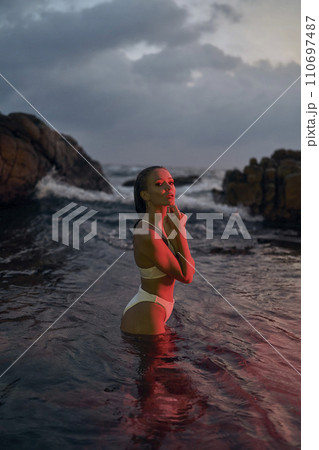 Attractive female enjoys nocturnal swim in sea under red illumination. Young woman in bikini experiences tranquil night dip amidst marine ambiance. Oceanic holiday, leisure travel for relax. 110697487