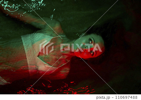 Scared woman floats in red-lit water at night, terrified expression, lagoon swimming, psychological themes of calm, dual self, introspection. Female frightened by unknown thoughts. 110697488