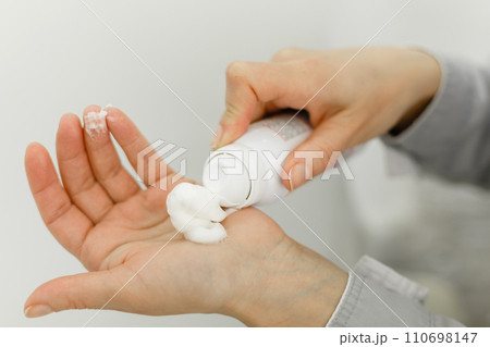 Hands of a pedicure master in a bathrobe and black gloves squeeze cream from the bottle. 110698147