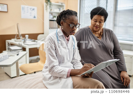 Medium full shot of Black woman physician in glasses holding clipboard and filling in medical record of senior female patient sitting in modern clinic office 110698446