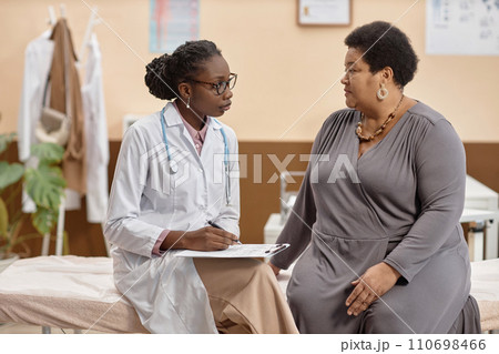 Medium full shot African American woman physician sitting on examination table holding clipboard while listening to mature female patients complaints 110698466