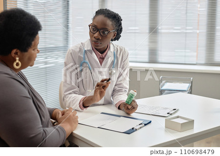 Young African American general practitioner explaining medication administration to female patient while sitting at table in hospital Young African American general practitioner explaining medication administration to female patient while sitting at table in hospital 110698479