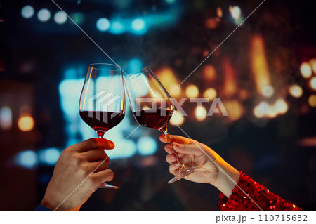 Anniversary celebration and meeting on a date. Male and female hands clinking glasses with red wine over restaurant background Anniversary celebration and meeting on a date. Male and female hands clinking glasses with red wine over restaurant background 110715632