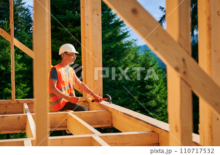 Carpenter constructing wooden frame two-story house near the forest. Man in glasses holding hammer, dressed in protective helmet and orange safety vest. Concept of ecological modern construction. Carpenter constructing wooden frame two-story house near the forest. Man in glasses holding hammer, dressed in protective helmet and orange safety vest. Concept of ecological modern construction. 110717532