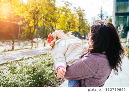 Cute lovely positive brunette mother carrying her happy disabilities daughter. Woman and child enjoy walk autumn sunny day outside. Sun glare effect. 110719645
