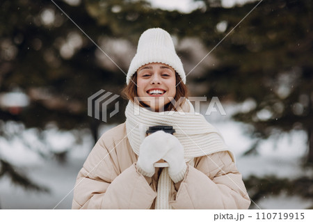 Happy smiling young woman with coffee hot drink cup enjoys winter weather at snowy winter park Happy smiling young woman with coffee hot drink cup enjoys winter weather at snowy winter park 110719915