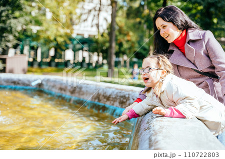 Happy family moments concept. Carful Caucasianmother and little girl child have a fun, enjoy, play with water of fountain outdoor in sunny park. 110722803