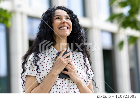 Optimistic young Latin American woman with curly hair smiling outside an office building, reflecting confidence and success. 110723005