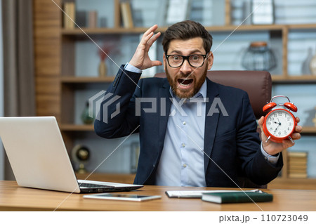 Surprised mature businessman in a suit holds a red alarm clock, expressing urgency, deadline stress in home office. 110723049