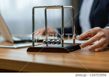 Mature businessman in a suit interacts with a Newton's cradle on his desk, symbolizing decision making and focus. 110723056