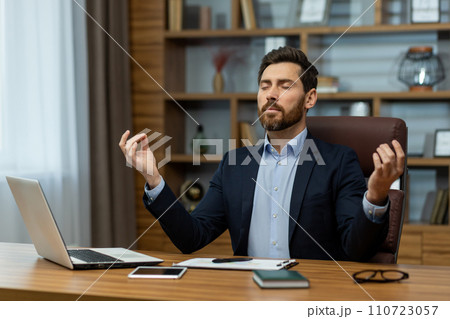 Experienced male professional in a suit practicing mindfulness meditation at his workplace for relaxation. 110723057