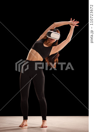 Portrait of young woman in fitness wear doing back exercises in virtual reality glasses against back studio background. 110723672