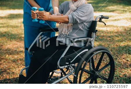 Young nurse or physiotherapist in scrubs helping a happy retired old woman do fitness exercises with light weight dumbbells at home. Concept of physiotherapy for seniors 110725597
