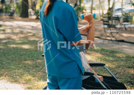 Young nurse or physiotherapist in scrubs helping a happy retired old woman do fitness exercises with light weight dumbbells at home. Concept of physiotherapy for seniors 110725598