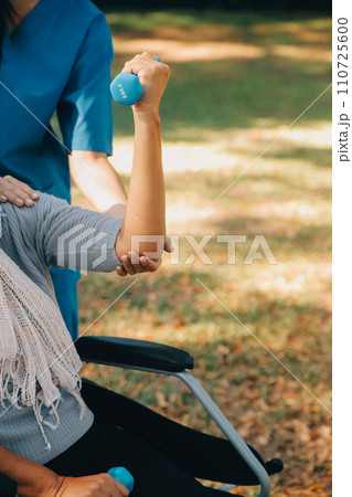 Young nurse or physiotherapist in scrubs helping a happy retired old woman do fitness exercises with light weight dumbbells at home. Concept of physiotherapy for seniors 110725600