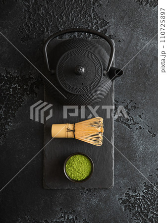 Green matcha tea powder with whisk and iron kettle on stone board on black background. Green matcha tea powder with whisk and iron kettle on stone board on black background. 110725897