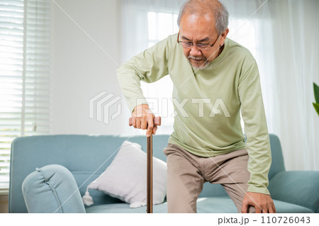 Elderly man suffering from knee pain ache holding handle of cane, senior old man typing to stand up from sofa alone with walking cane stick to walk at home, retirement medical healthcare concept Elderly man suffering from knee pain ache holding handle of cane, senior old man typing to stand up from sofa alone with walking cane stick to walk at home, retirement medical healthcare concept 110726043