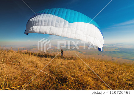 A paraglider takes off from a mountainside with a blue and white canopy and the sun behind. A paraglider is a silhouette. The glider is sharp, with little wing movement. 110726465