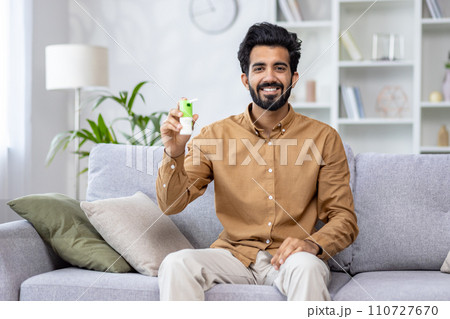 Portrait of young Indian man sitting on sofa at home, holding and showing throat spray. Smiling and looking at the camera. 110727670