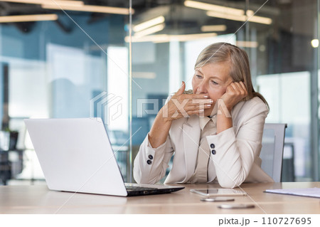 Tired senior woman sitting at desk in front of laptop in office in business suit, head on hand and yawning while covering mouth with hand. Tired senior woman sitting at desk in front of laptop in office in business suit, head on hand and yawning while covering mouth with hand. 110727695