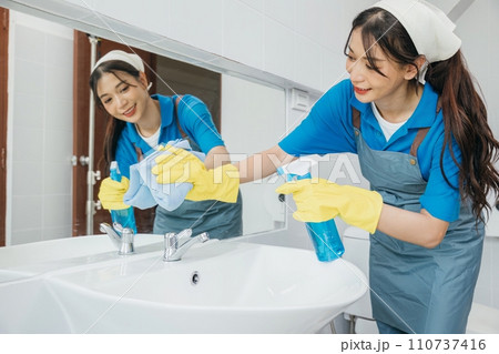 A cheerful maid in rubber gloves wipes bathroom mirror with a rag ensuring cleanliness and shining reflection. Hotel service emphasizes professional cleaning for hygiene. Maid cleaning at home 110737416