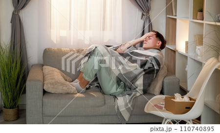 Sickness fever. Seasonal flu. Ill tired man wrapped in blanket laying on couch suffering from bad headache in living room with drugs at home. 110740895