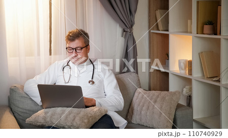 Work leisure. Doctor break. Online communication. Relaxed healthcare specialist man in white uniform typing on laptop sitting on couch in cozy interior. 110740929