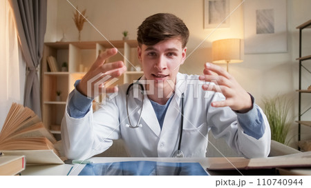 Young doctor. Healthcare specialist. Online consultation. Positive man in medical uniform talking at video appointment explaining diagnosis sitting at desk in clinic. Young doctor. Healthcare specialist. Online consultation. Positive man in medical uniform talking at video appointment explaining diagnosis sitting at desk in clinic. 110740944