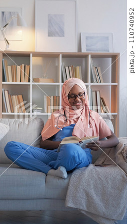 Doctor study. Medical education. Smiling woman student in hijab reading nurse course relaxing on sofa at hospital staff room. Doctor study. Medical education. Smiling woman student in hijab reading nurse course relaxing on sofa at hospital staff room. 110740952