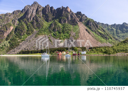 Sailboats glide past a red Rorbu against green cliffs in Norway Sailboats glide past a red Rorbu against green cliffs in Norway 110745937