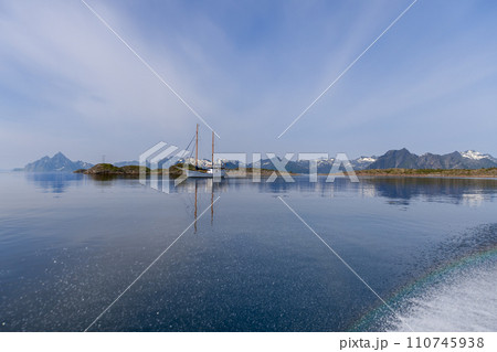 Calm sea mirrors a sailboat and mountains with a faint rainbow in Lofoten 110745938